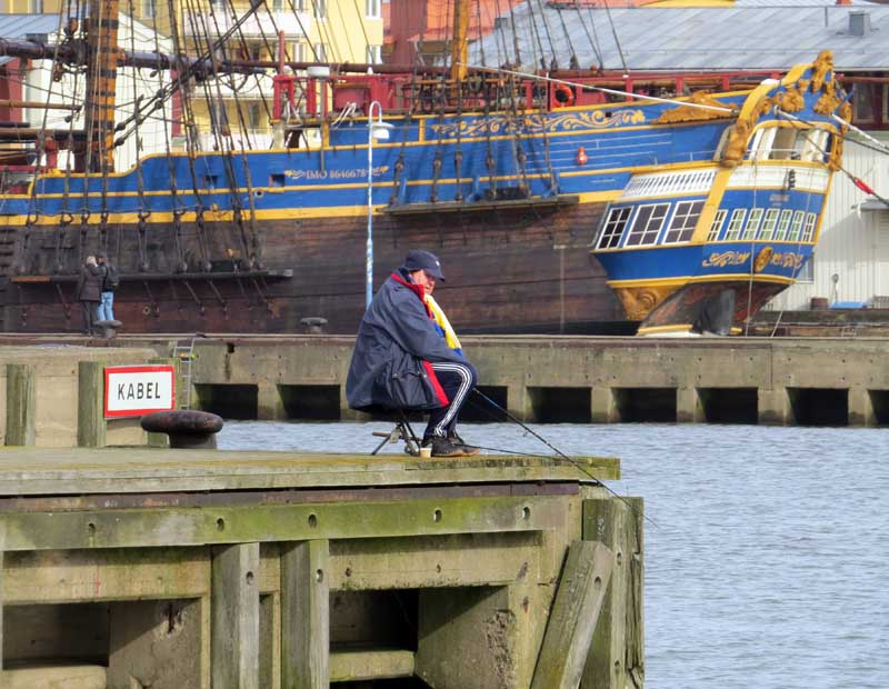 Man in blue and yellow scarf angling off a pier with the replica East Indiaman Götheborg behind