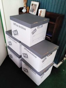 A stack of archive boxes fill of books standing in front of one of my mother's now empty bookshelves