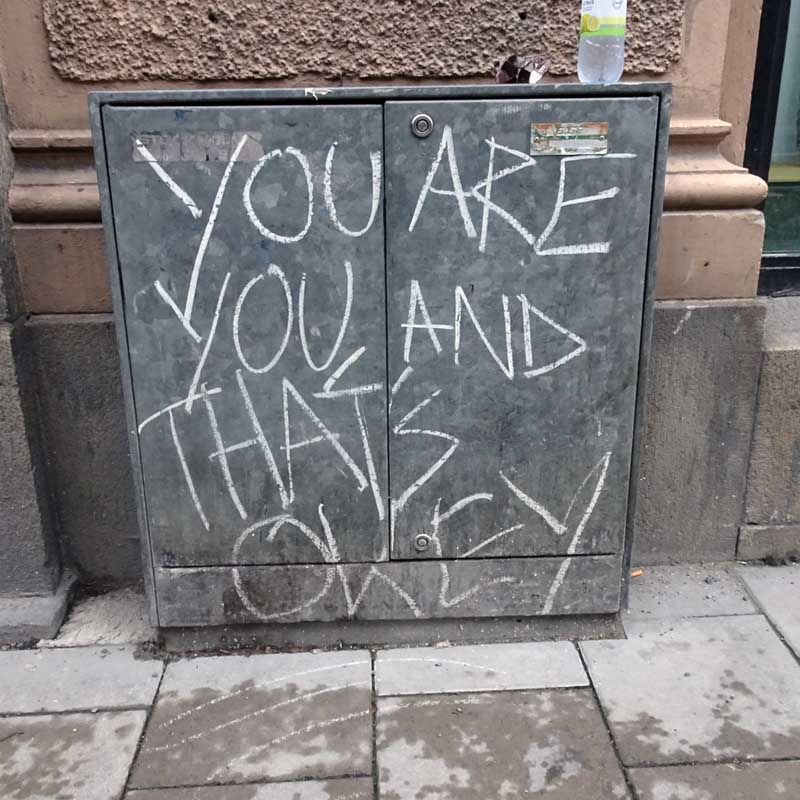 A chalked graffiti statement on a grey metal electrical cabinet in Stockholm reads: You are you and that's okey