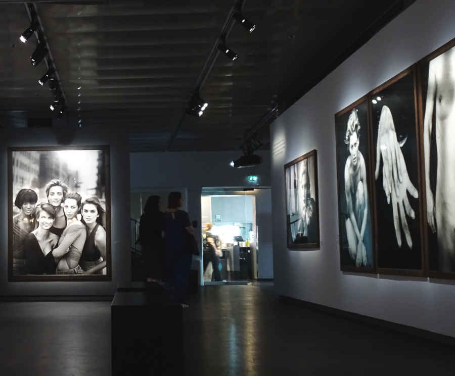 Second series of monumental black-and-white photographs by Peter Lindbergh in an exhibition hall at Stockholm's Fotografiska gallery. A couple of gallery goers are in shadow and the entrance to the museum's shop can be seen in the distance.